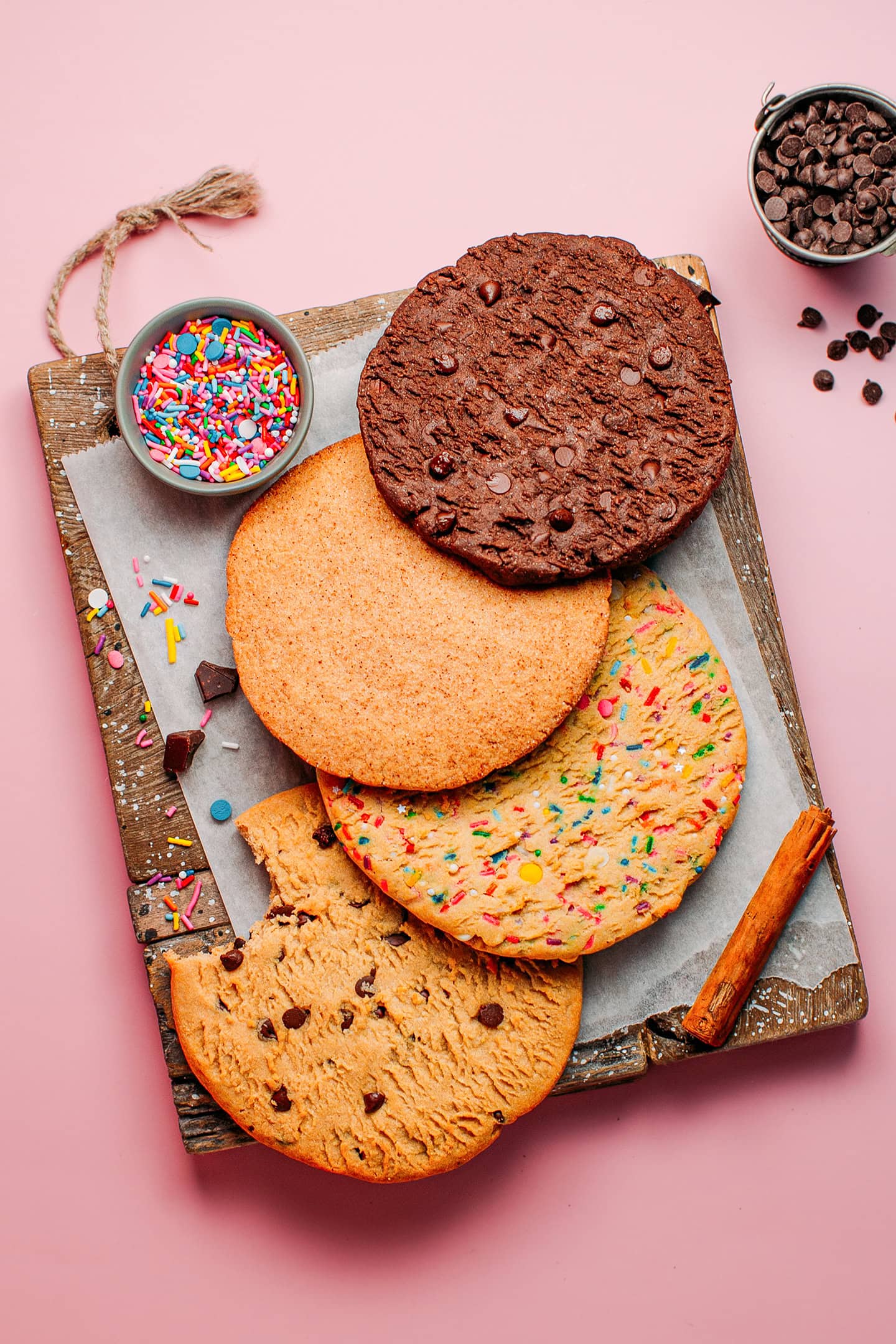 Four different cookies with chocolate chips, sprinkles, cinnamon sugar, and chocolate, on a wooden board.