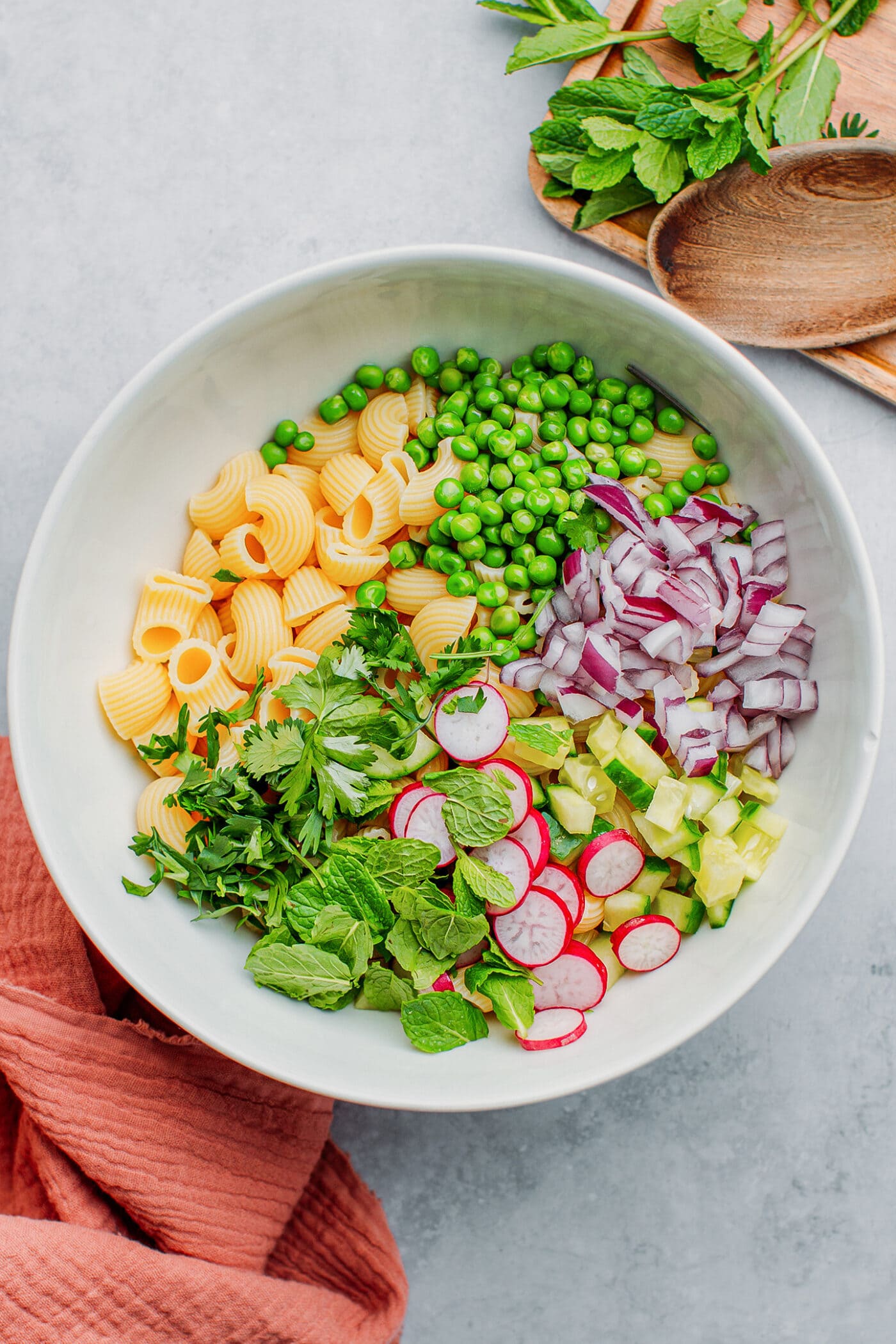 Cooked macaroni, red onions, radishes, cucumber, and fresh herbs in a bowl.