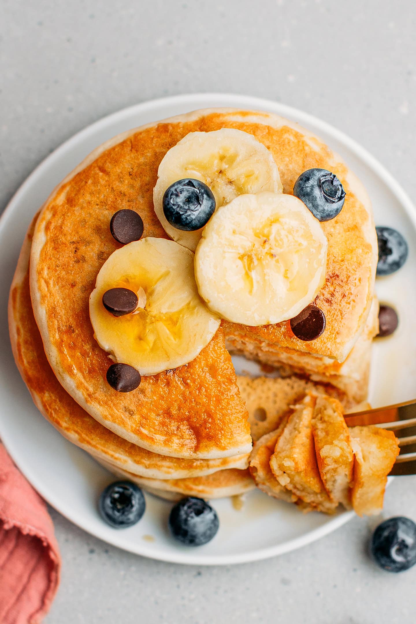 Top view of a atack of vegan pancakes drizzled with maple syrup and topped with banana slices, blueberries, and chocolate chips.