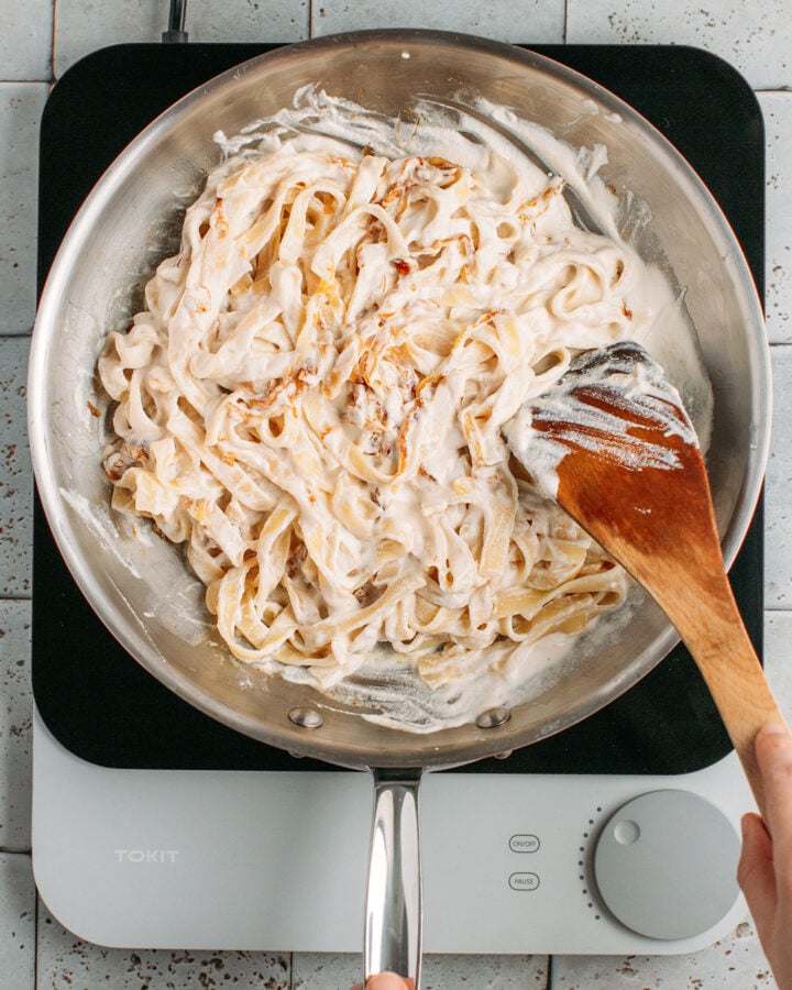 Tossing pasta with caramelized onions and cashew cream in a skillet.