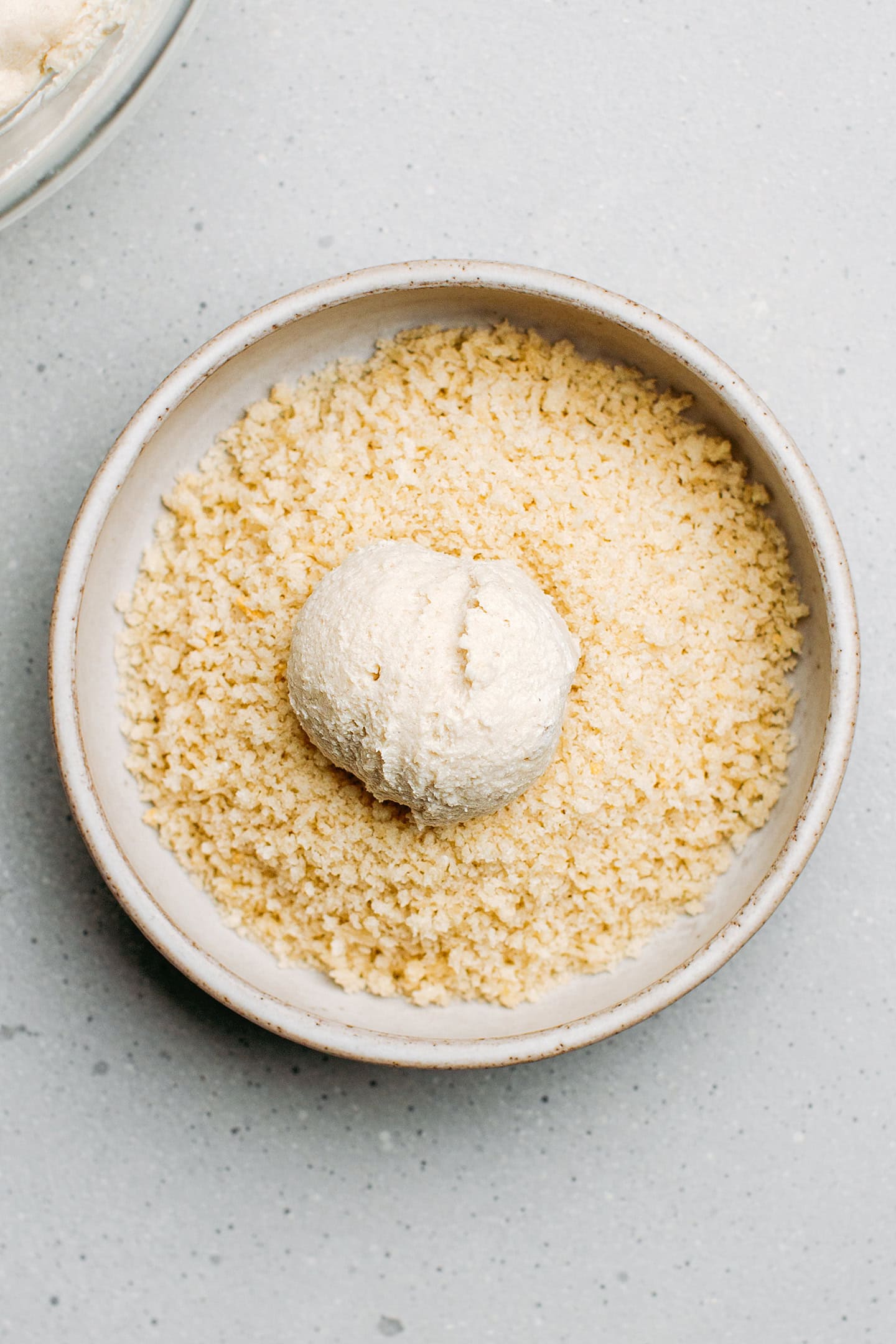 Vegan cheese ball in a bowl containing breadcrumbs.