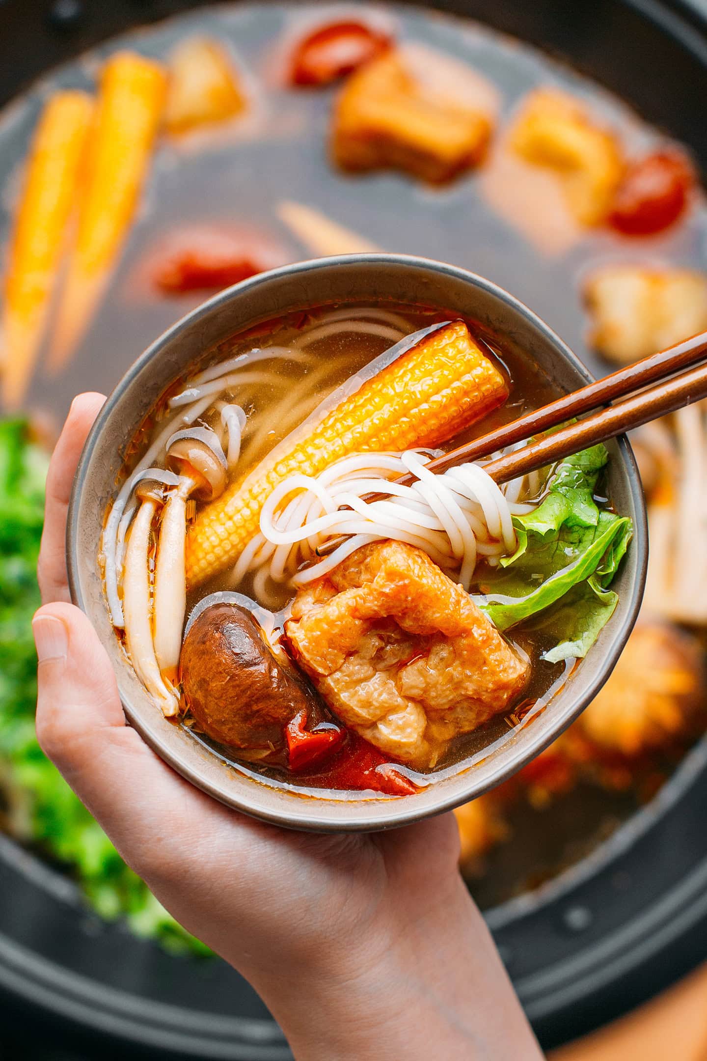 Holding a bowl filled with rice noodles, baby corn, tofu, and mushrooms.