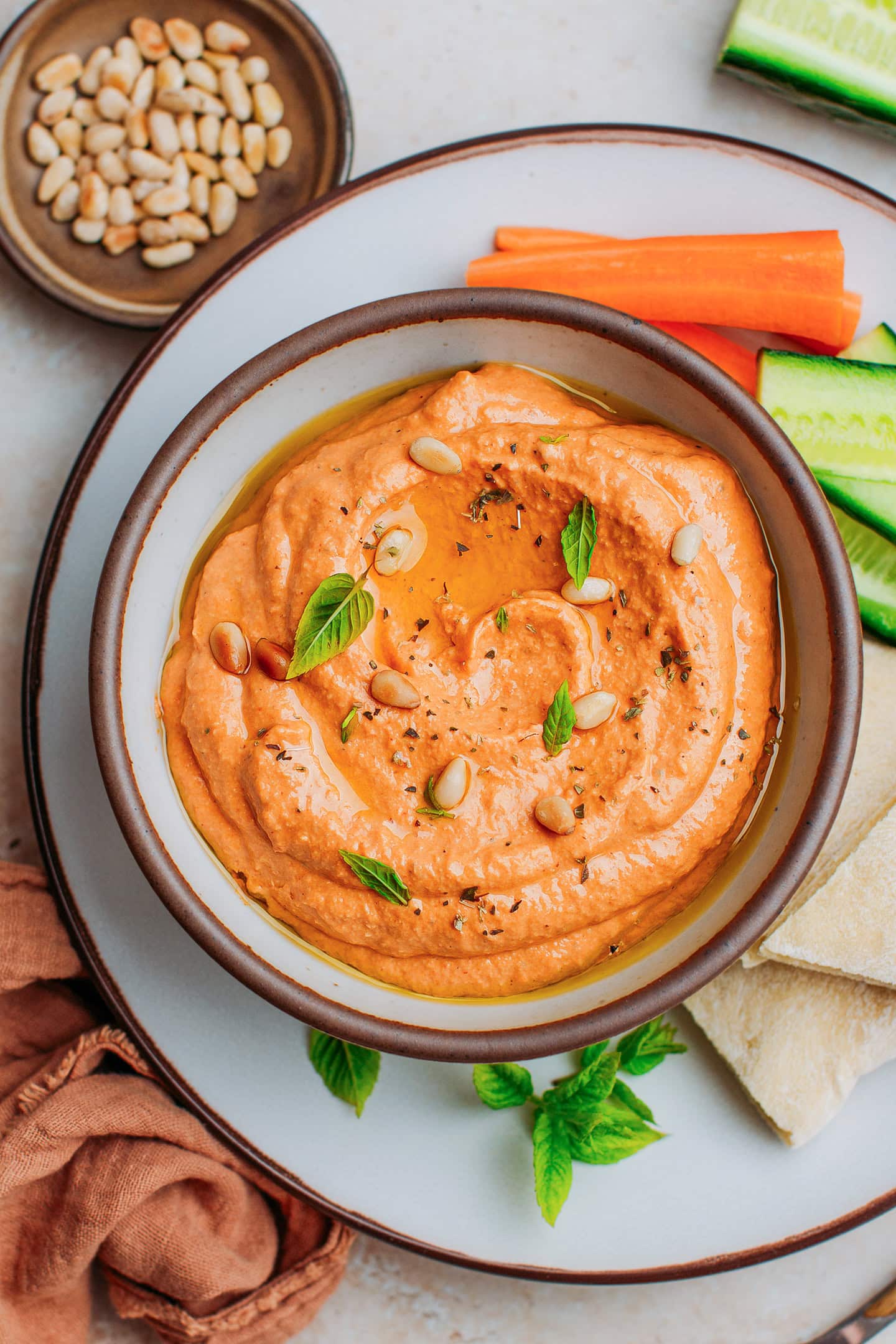 Top view of a red bell pepper dip topped with pine nuts, mint leaves, and oregano.