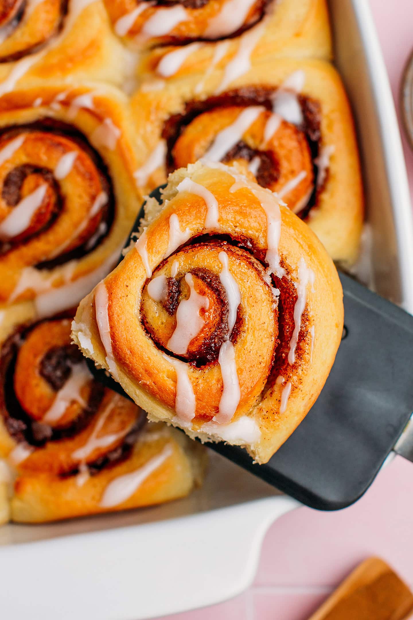 Holding a cinnamon roll using a spatula over a baking dish.