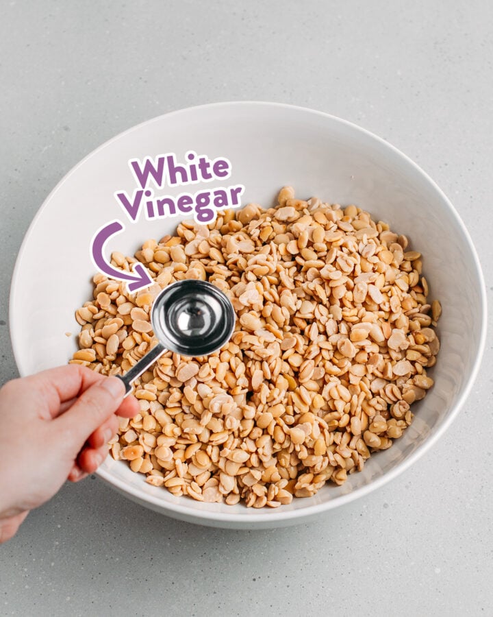 Holding a tablespoon of white vinegar over a bowl filled with cooked soybeans.