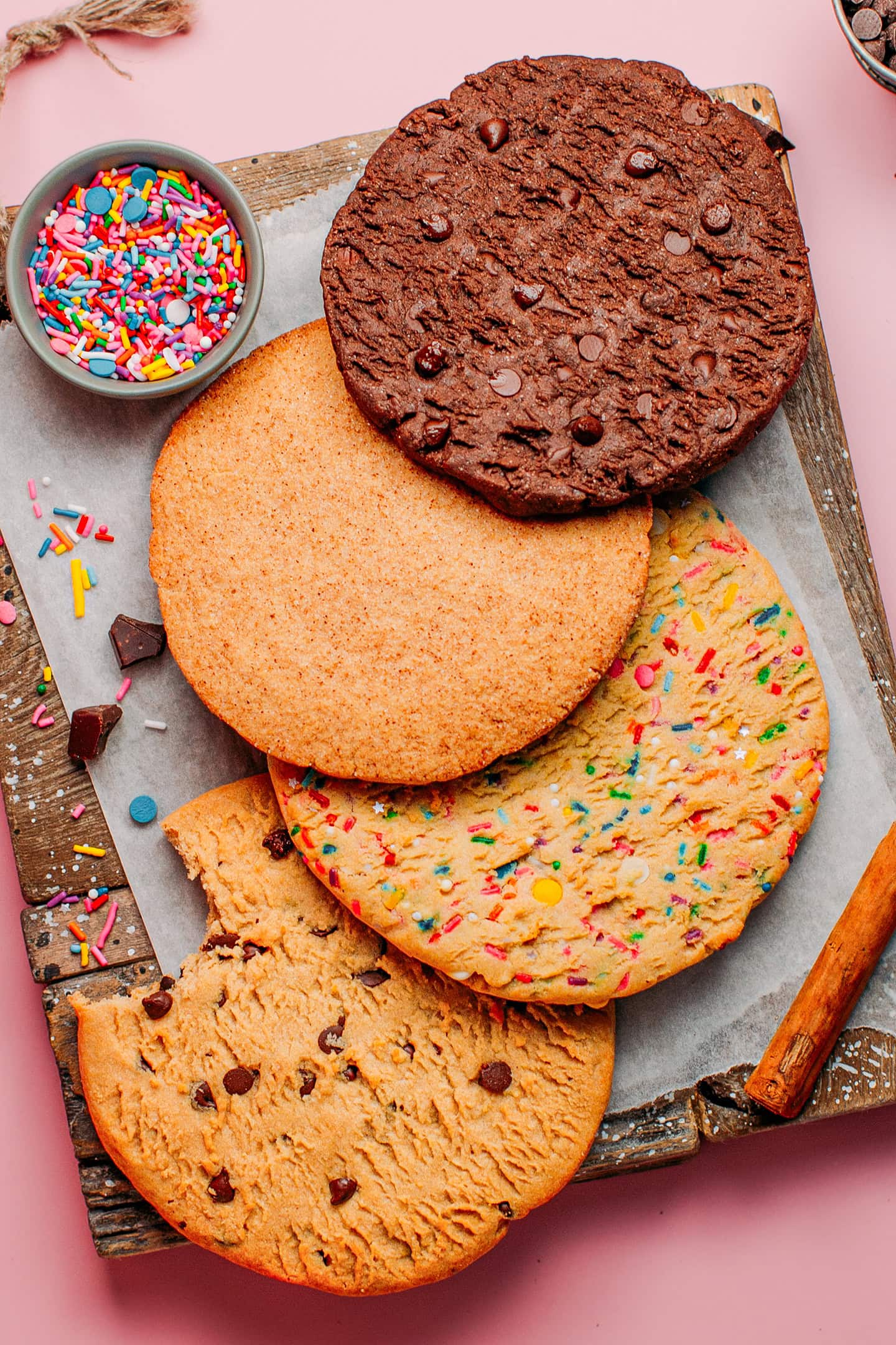 Four different cookies with chocolate chips, sprinkles, cinnamon sugar, and chocolate, on a wooden board.