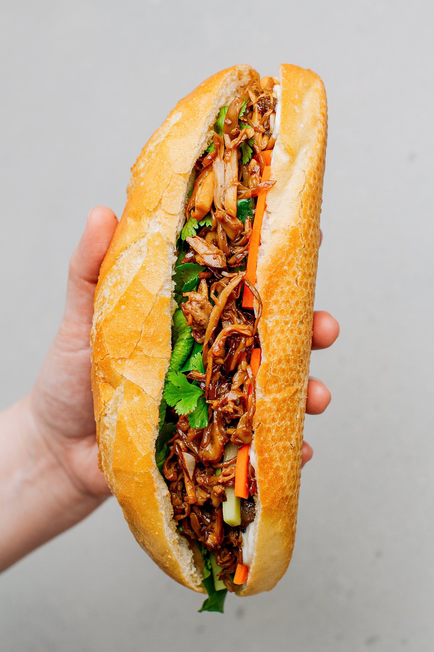 Holding a banh mi baguette filled with pulled mushrooms, lettuce, carrots, and cucumber.