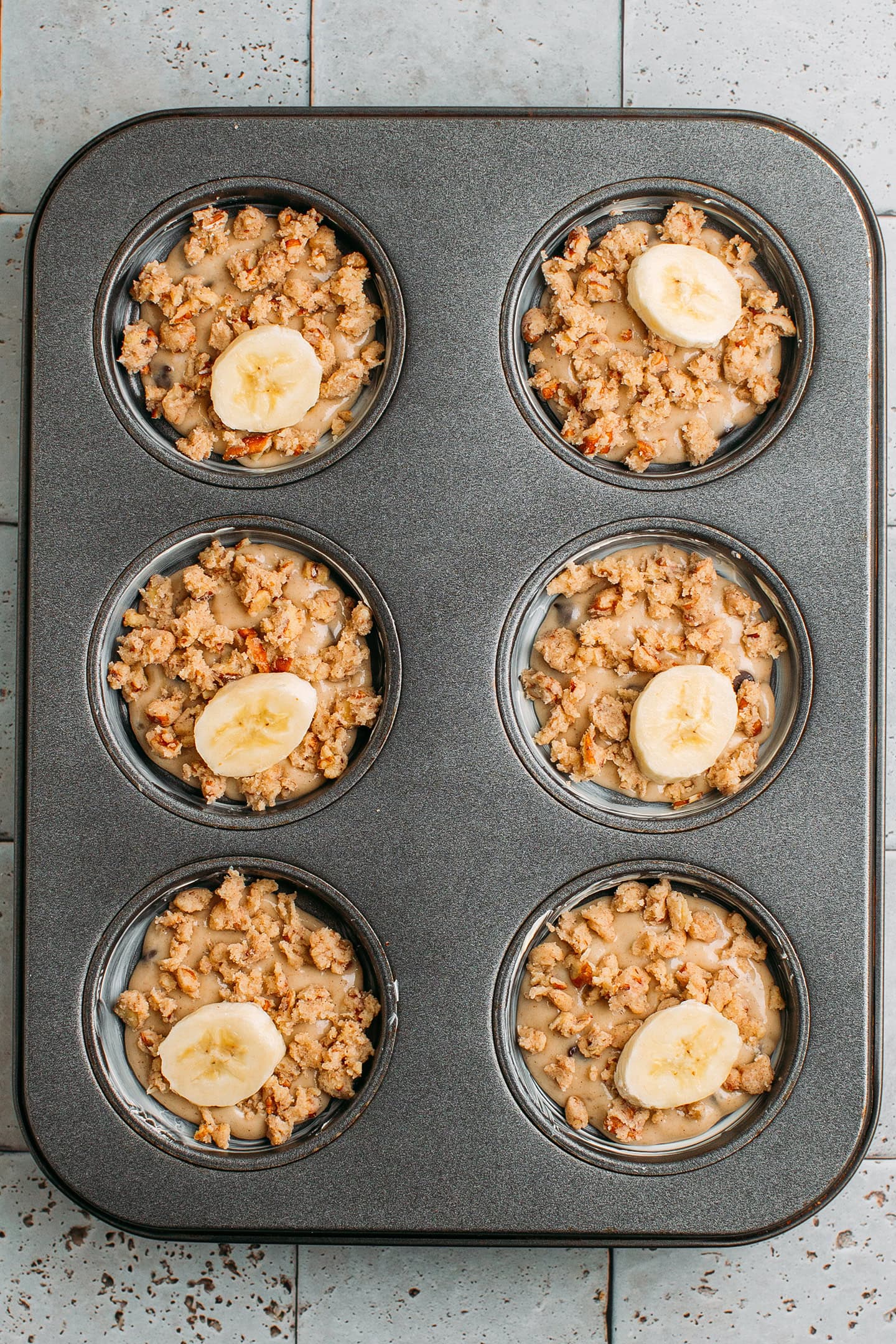 Muffin pan filled with batter and topped with streusel topping and banana slices.
