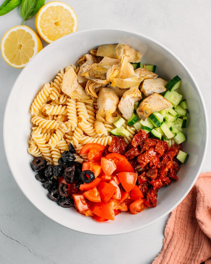 Cooked pasta, artichokes, olives, tomatoes, and cucumber in a mixing bowl.