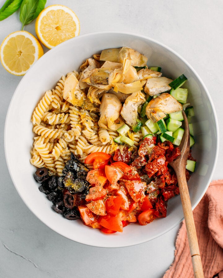Cooked fusilli, diced cucumber, dried tomatoes, artichokes, and vinaigrette in a mixing bowl.