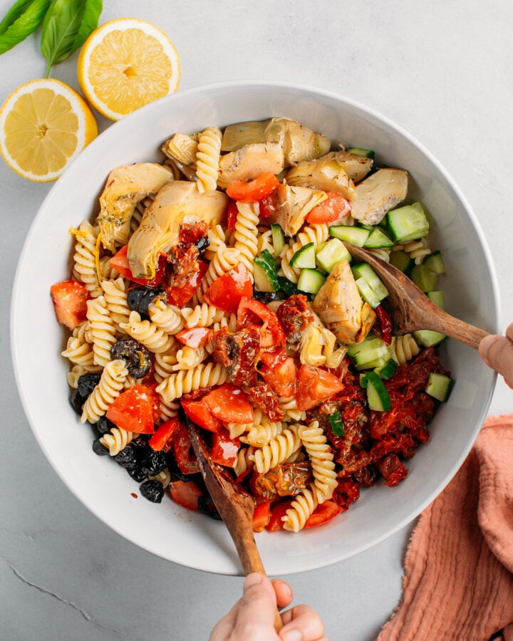 Tossing cooked pasta with vegetables and vinaigrette in a mixing bowl.