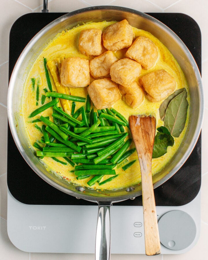 Green beans, tofu puffs, and bay leaves with coconut milk in a skillet.