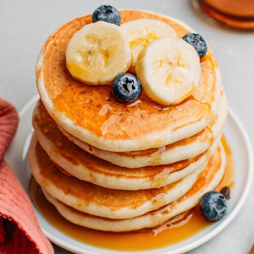Stack of vegan pancakes drizzled with maple syrup and topped with banana slices and blueberries.