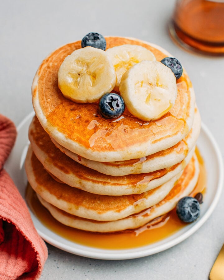 Stack of vegan pancakes drizzled with maple syrup and topped with banana slices and blueberries.