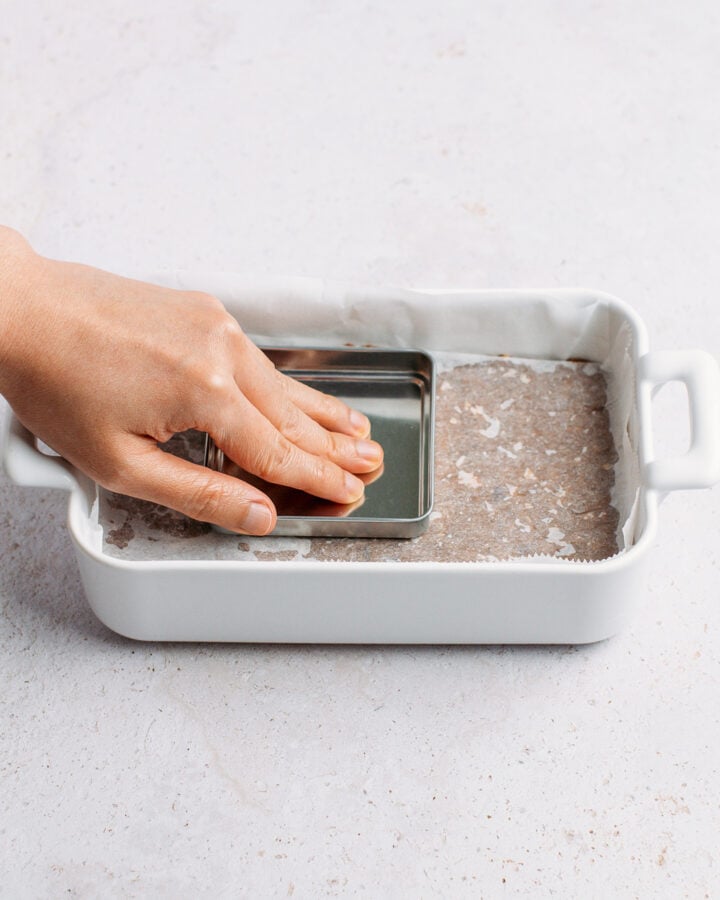 Using a metal cap to press down protein bar mixture firmly into an even layer inside a baking dish.