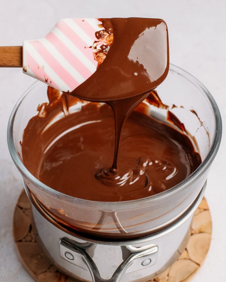Holding a spatula dipped in melted dark chocolate over a bowl of chocolate.