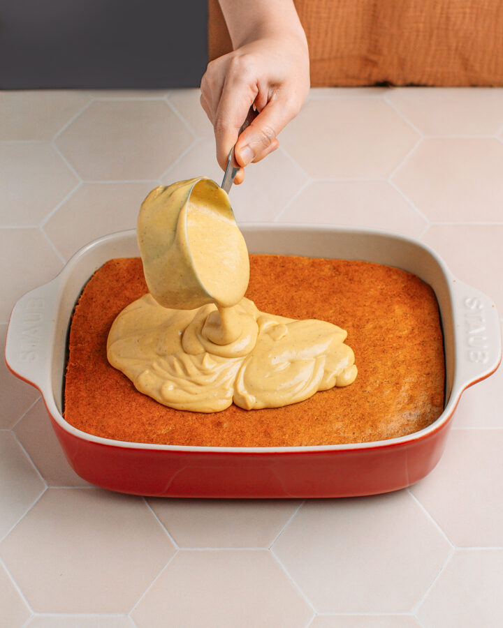 Pouring pumpkin cream over a pumpkin sponge cake layer inside a baking dish.