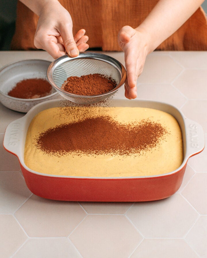 Dusting cocoa powder over pumpkin cream inside a baking dish.