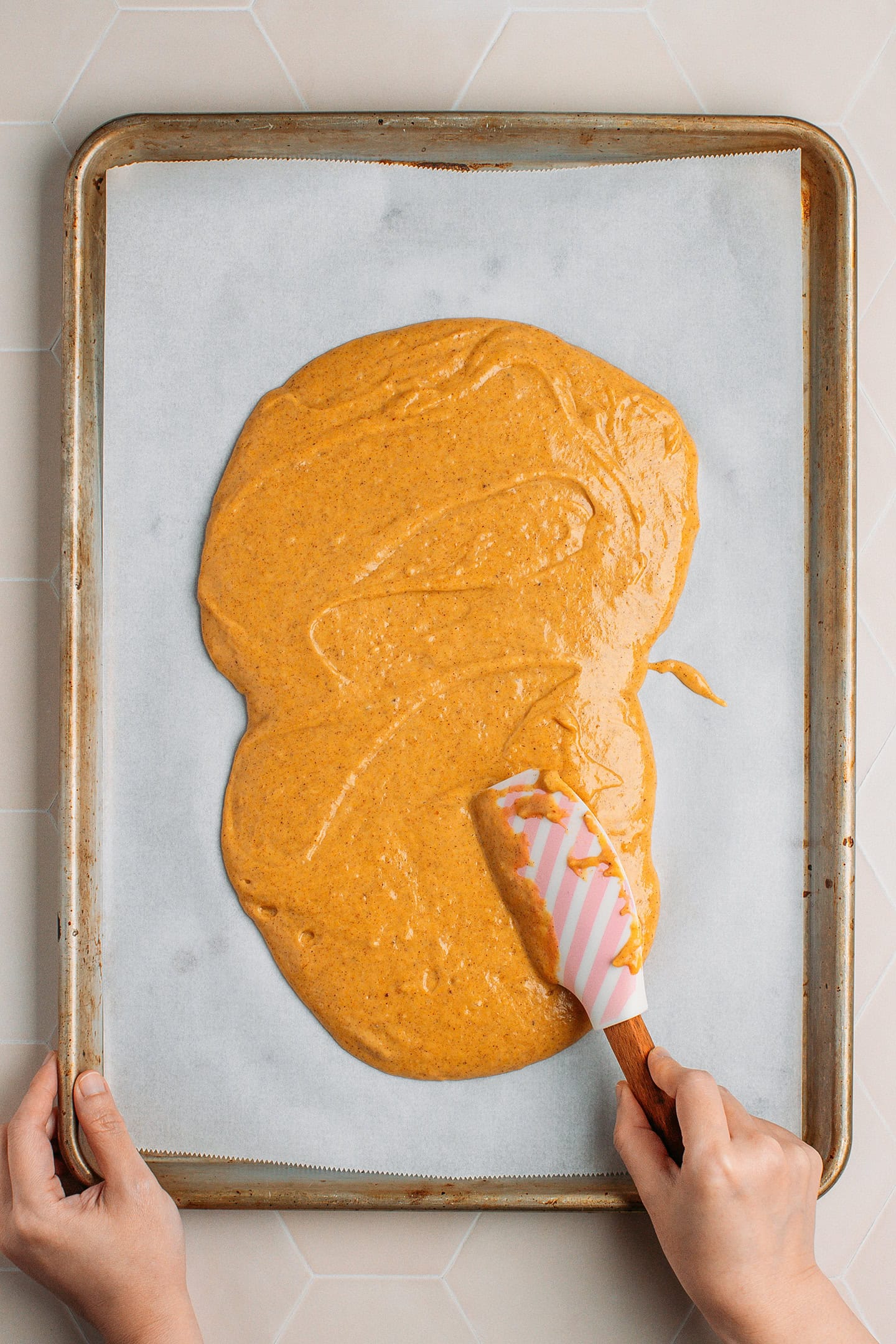 Using a spatula to spread pumpkin cake batter on a baking sheet.