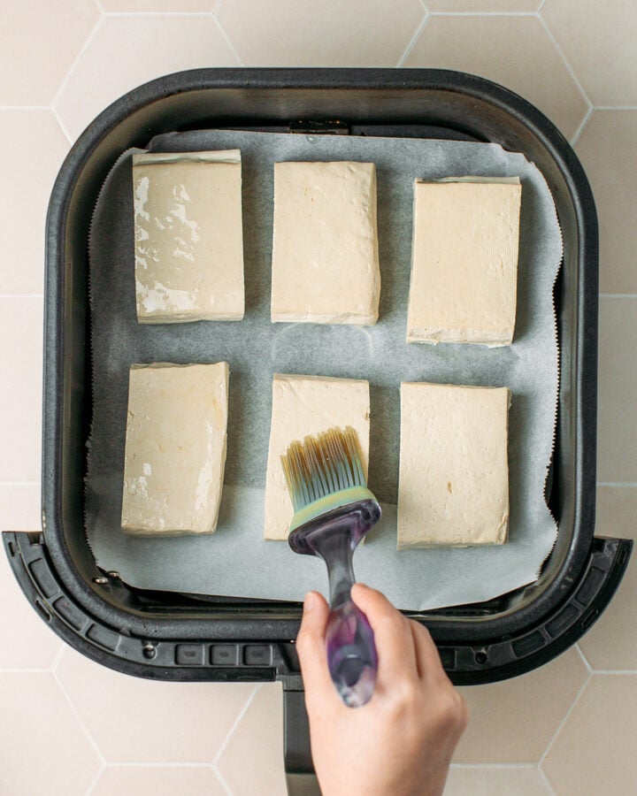 Brushing oil over firm tofu placed in an air fryer basket.