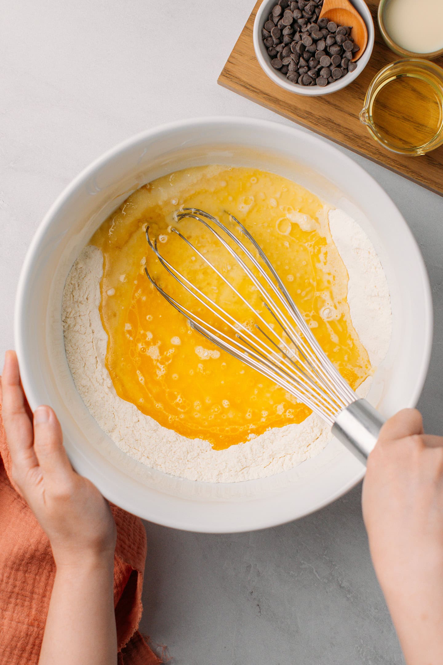 Whisking melted butter, milk, and oil with flour and sugar in a mixing bowl.