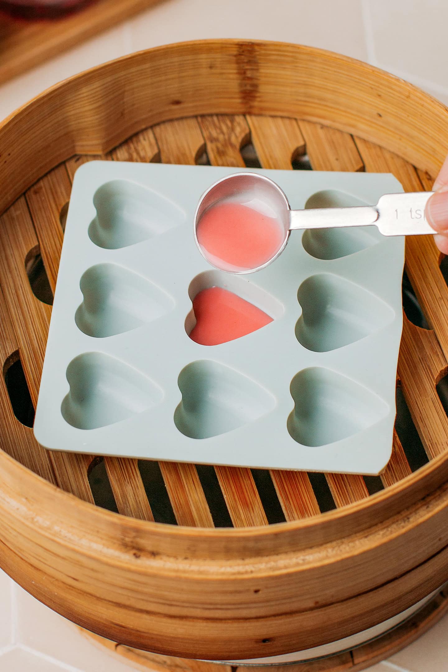 Using a teaspoon to pour a red batter into a heart-shaped silicone mold.