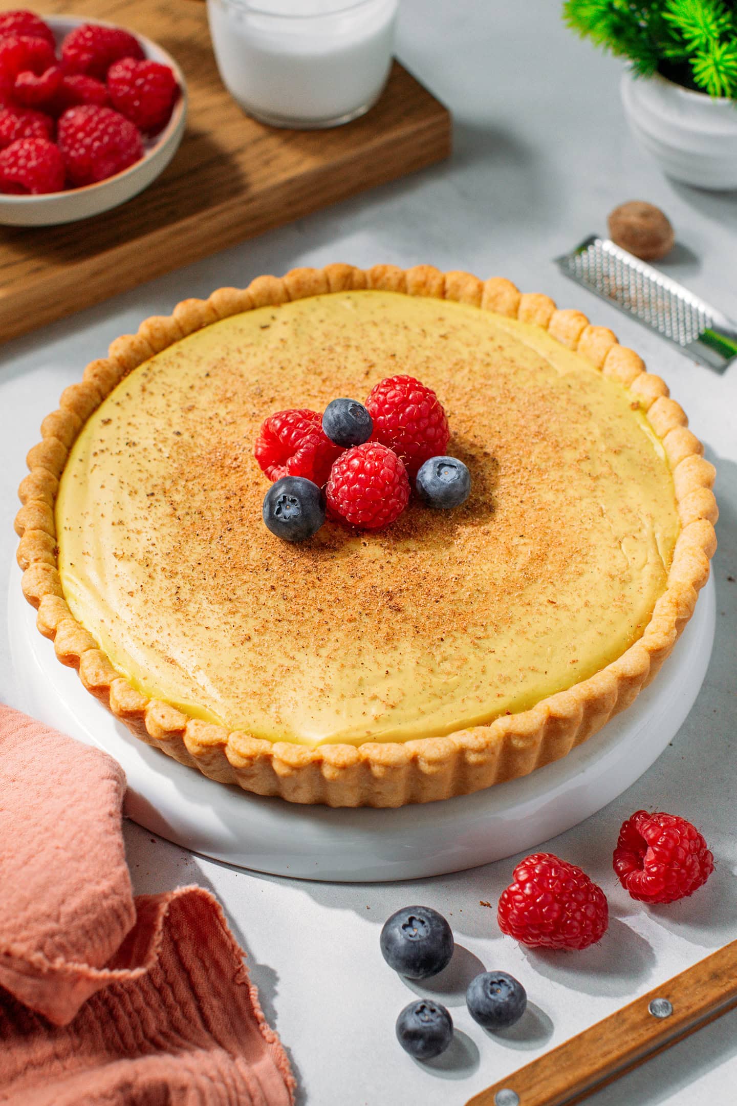 Whole custard tart topped with ground nutmeg, raspberries, and blueberries on a white plate.