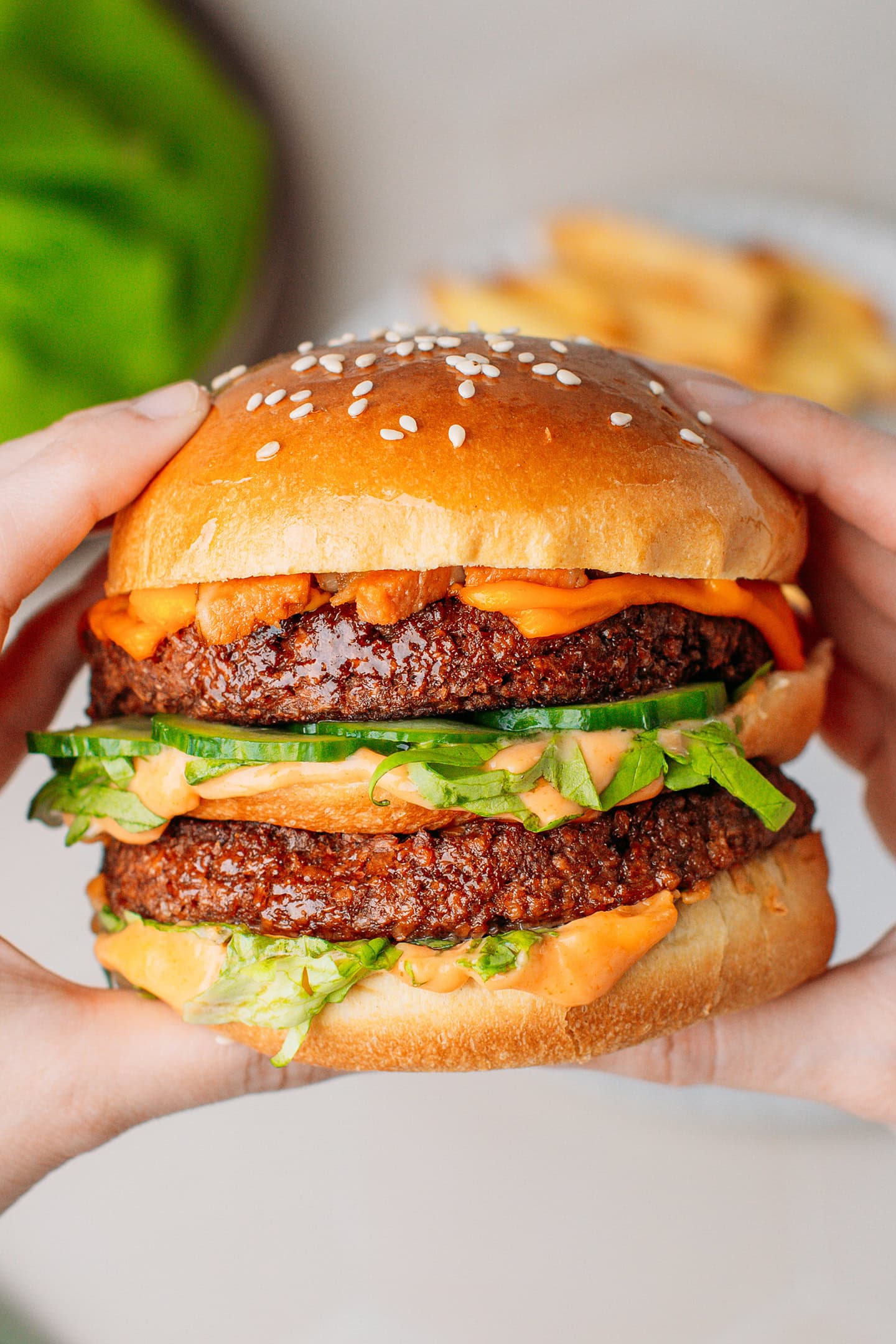 Holding a homemade hamburger stuffed with vegan meat patties, cucumbers, and vegan cheese.