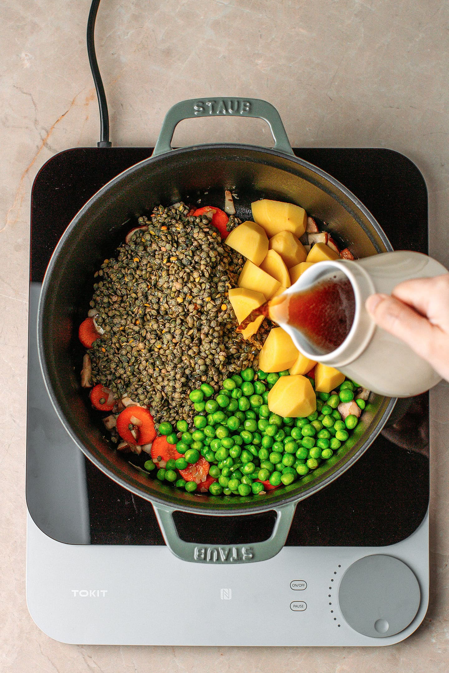 Pouring beer into a pot containing green lentils, diced potatoes, green peas, and carrots.